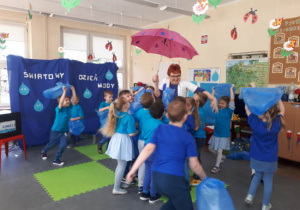 Dzieci biegające z niebieskimi workami foliowymi w górze podczas zabawy ruchowej. Nauczycielka stojąca po środku z parasolem rozłożonym nad głową.